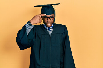 Young african american man wearing graduation cap and ceremony robe pointing unhappy to pimple on...