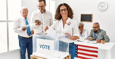 Middle age american voter woman smiling happy putting ballot in voting box at vote center.