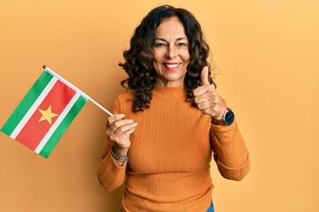Middle age hispanic woman holding suriname flag smiling happy and positive, thumb up doing...