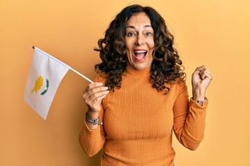 Middle age hispanic woman holding cyprus flag screaming proud, celebrating victory and success very excited with raised arm