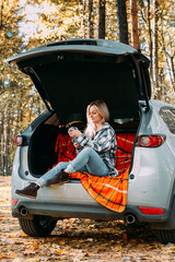 a blonde adult girl in a plaid shirt is drinking coffee sitting in the open trunk of a car. picnic in the car.