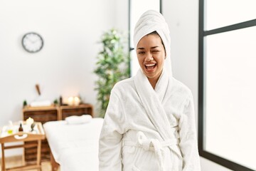 Young brunette woman wearing towel and bathrobe standing at beauty center winking looking at the...