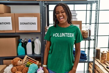 Young african american woman working wearing volunteer t shirt at donations stand winking looking...