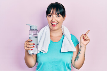 Young hispanic woman wearing sportswear holding water bottle smiling happy pointing with hand and finger to the side