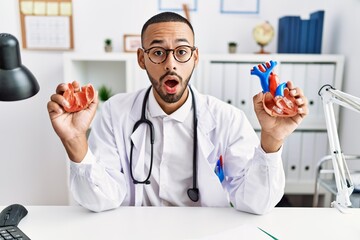 African american doctor man holding anatomical heart at the clinic in shock face, looking skeptical...