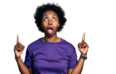 African american woman with afro hair wearing casual purple t shirt amazed and surprised looking up and pointing with fingers and raised arms.