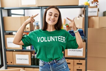 Young brunette woman wearing volunteer t shirt at donations stand smiling pointing to head with...