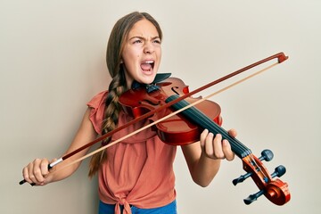 Beautiful brunette little girl playing violin angry and mad screaming frustrated and furious,...