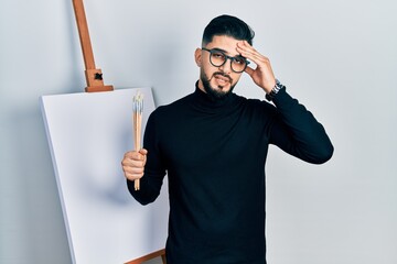 Handsome man with beard holding brushes close to easel stand worried and stressed about a problem...