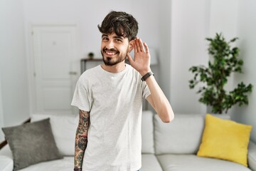 Hispanic man with beard at the living room at home smiling with hand over ear listening an hearing to rumor or gossip. deafness concept.