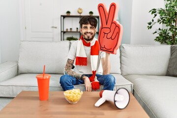 Naklejka premium Hispanic man with beard supporting football team at home sitting on the sofa looking positive and happy standing and smiling with a confident smile showing teeth