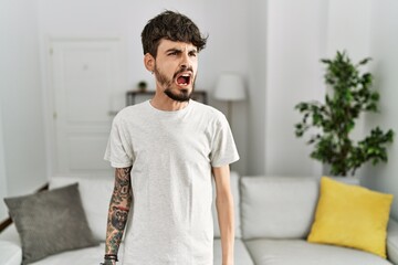 Hispanic man with beard at the living room at home angry and mad screaming frustrated and furious, shouting with anger. rage and aggressive concept.