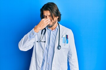 Middle age handsome man wearing doctor uniform and stethoscope smelling something stinky and disgusting, intolerable smell, holding breath with fingers on nose. bad smell