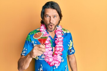 Middle age handsome man wearing hawaiian lei drinking cocktail scared and amazed with open mouth...