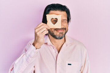 Middle age caucasian man holding bread loaf with heart shape looking positive and happy standing and smiling with a confident smile showing teeth