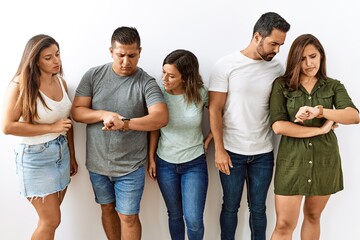 Group of young hispanic friends standing together over isolated background checking the time on wrist watch, relaxed and confident