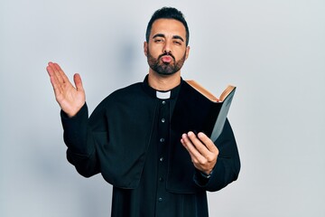 Handsome hispanic priest man with beard holding bible and christian cross looking at the camera...