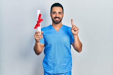 Handsome hispanic man with beard wearing blue male nurse uniform holding diploma smiling with an...