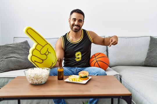 Young hispanic man with beard holding basketball ball cheering tv game smiling happy pointing with hand and finger