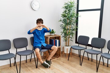 Fototapeta premium Young hispanic man sitting at doctor waiting room with neck injury smiling in love showing heart symbol and shape with hands. romantic concept.