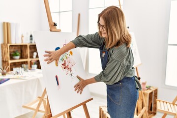 Middle age artist woman smiling happy painting canvas with painted hands at art studio.