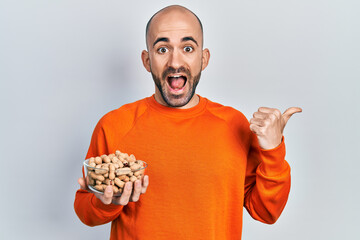 Young bald man holding peanuts pointing thumb up to the side smiling happy with open mouth