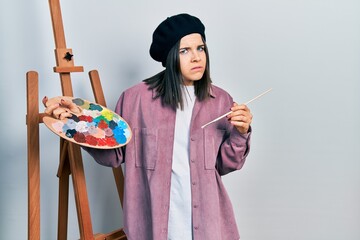 Young brunette woman standing by empty easel stand holding palette clueless and confused expression. doubt concept.