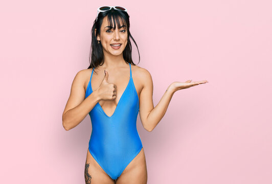 Young hispanic woman wearing swimsuit and sunglasses showing palm hand and doing ok gesture with thumbs up, smiling happy and cheerful