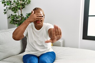 Mature hispanic woman sitting on the sofa at home covering eyes with hands and doing stop gesture with sad and fear expression. embarrassed and negative concept.