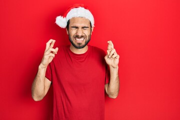Fototapeta na wymiar Young hispanic man wearing christmas hat gesturing finger crossed smiling with hope and eyes closed. luck and superstitious concept.