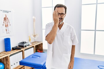Middle age man with beard working at pain recovery clinic covering one eye with hand, confident smile on face and surprise emotion.