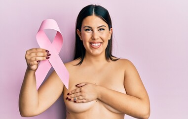 Beautiful brunette woman holding pink cancer ribbon covering bust smiling and laughing hard out...