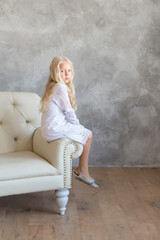 Fototapeta na wymiar Little blonde princess in a white casual dress sits on a white leather sofa against a gray wall. Fashion for kids concept