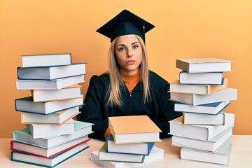 Young caucasian woman wearing graduation ceremony robe sitting on the table looking sleepy and tired, exhausted for fatigue and hangover, lazy eyes in the morning.