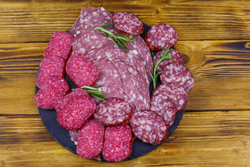 Different tasty sliced salami sausage on a black slate serving board on wooden table. Top view
