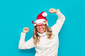 Fototapeta na wymiar A young smiling blonde woman in glasses with deer antlers, a Christmas white warm sweater, Santa hat is happy with her hands up isolated on a color blue background. Christmas and New Year concept