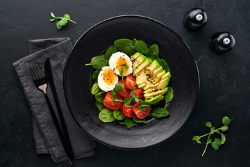 Avocado, cherry tomato, spinach and chicken egg, microgreens peas and black sesame seeds fresh salad in black bowl on black slate, stone or concrete background. Healthy food concept. Top view.
