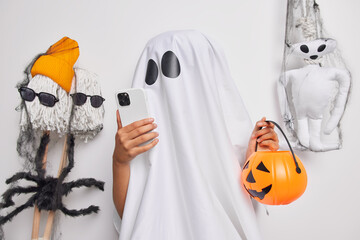 Spooky holiday concept. Faceless ghost covered with white sheet checks notification on smartphone...