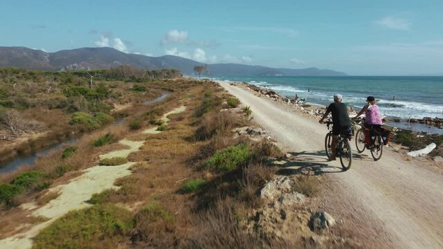 A married couple on their electric pedelec bikes, active and sporty seniors riding along the scenic seaside beach ocean in Italy on a warm summer day. Aerial drone of a healthy bicycle tour.