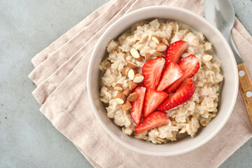 Oatmeal porridge with strawberry slices, nuts almonds and honey in blue bowl on grey table. Healthy eating, dieting, vegetarian food concept. Place for text