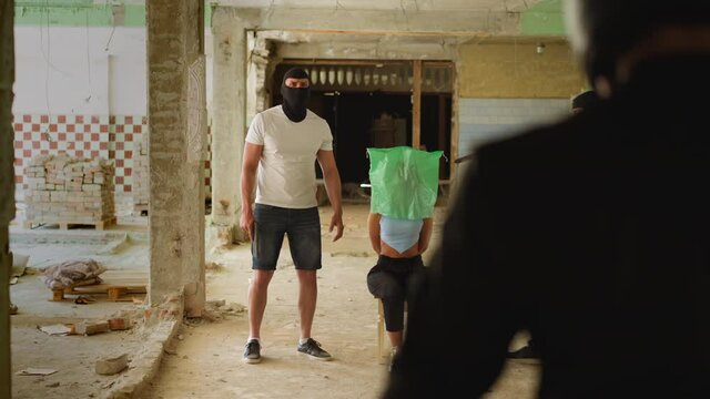Bandit in mask cocks pistol on woman hostage looking with companion at walking man saver in helmet in old abandoned building slow motion
