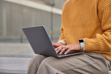 Cropped shot of unrecognizable woman works on new project keyboards information on laptop computer wears knitted yellow sweater and trousers. Anonymuous female model presses buttons creats text