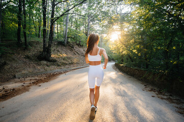 A young beautiful girl in white sports clothes is running with her back, on the road in a dense...