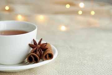 White cup of tea with cinnamon on a beige plaid. Christmas lights and copy space.