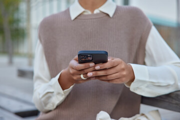 Cropped shot of unknown woman in elegant clothes holds mobile phone surfs wireless internet makes shopping online sits on bench outside connected to wireless internet communicates distantly.