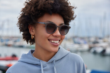 Close up shot of cheerful smiling beautiful woman with curly hair wears sunglasses and sweatshirt looks happily away poses against blurred harbor background strolls outside admires marine view