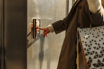 close-up of a finger presses the elevator button. a woman with shopping bags in a beige raincoat...