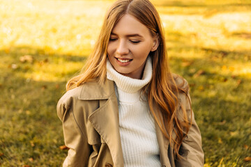 beautiful woman in a beige raincoat sits on the grass in the park, against the background of autumn leaf fall