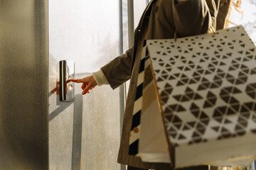 close-up of a finger presses the elevator button. a woman with shopping bags in a beige raincoat...