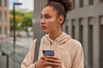 Fototapeta na wymiar Horizontal shot of thoughtful sporty girl in sweatshirt holds digital modern mobile phone sends text messages carries karemat poses in urban setting rests after cardio training. Healthy lifestyle